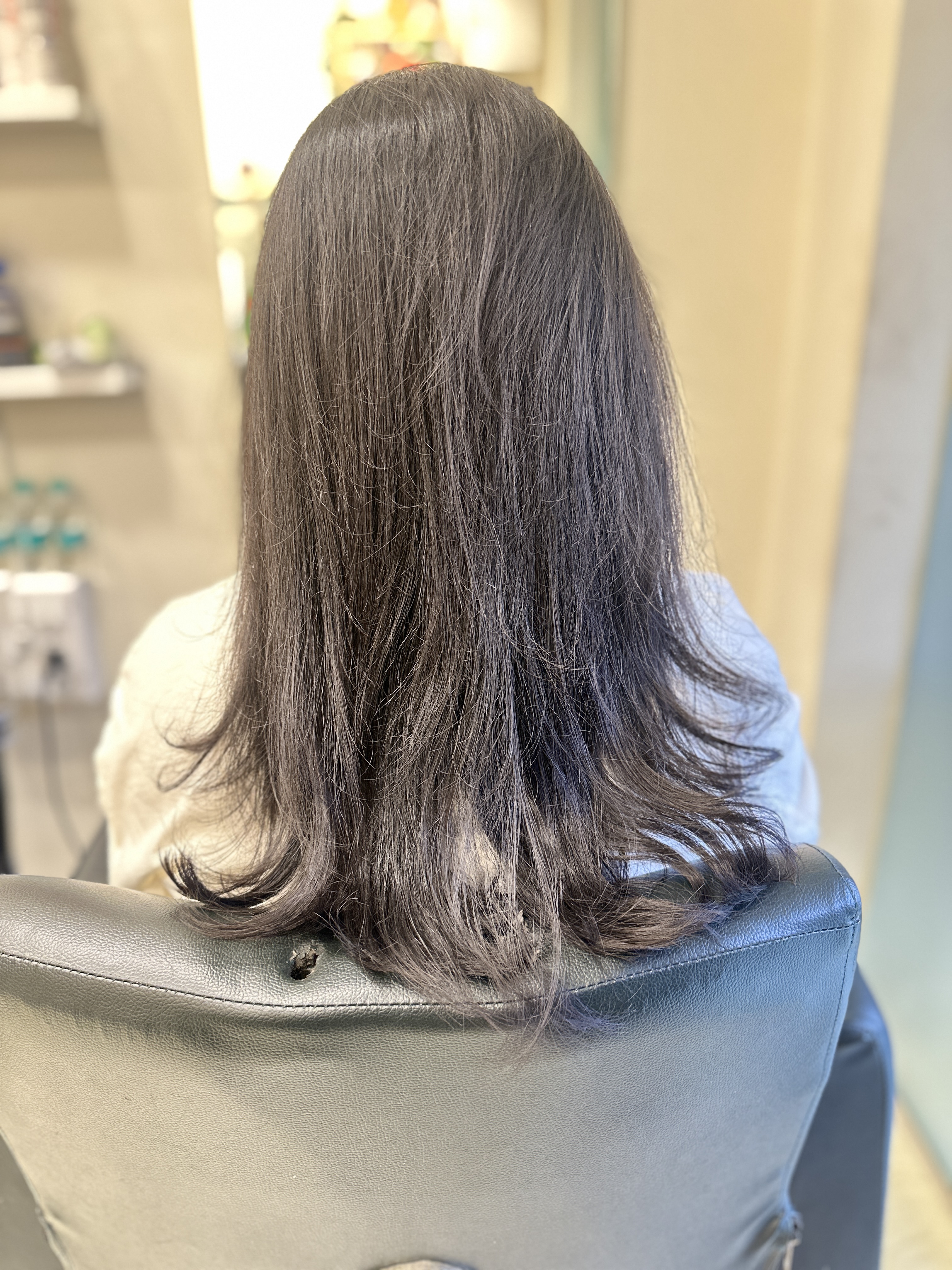 Back view of a woman with dark, smooth, healthy-looking medium-length hair, styled with soft waves at the ends, sitting in a salon chair at Seema's Hair & Skin Clinique.